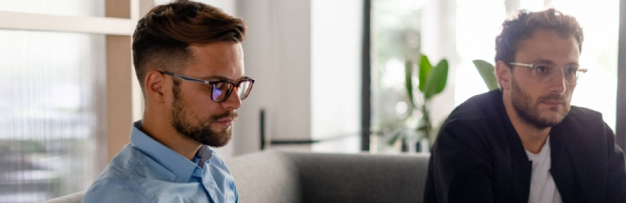 Two men wearing glasses are seated on a couch, engaged in conversation. 