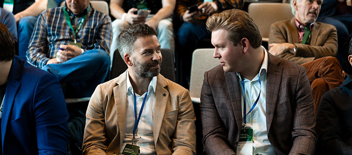 Two men engaged in conversation at a conference with attendees in the background