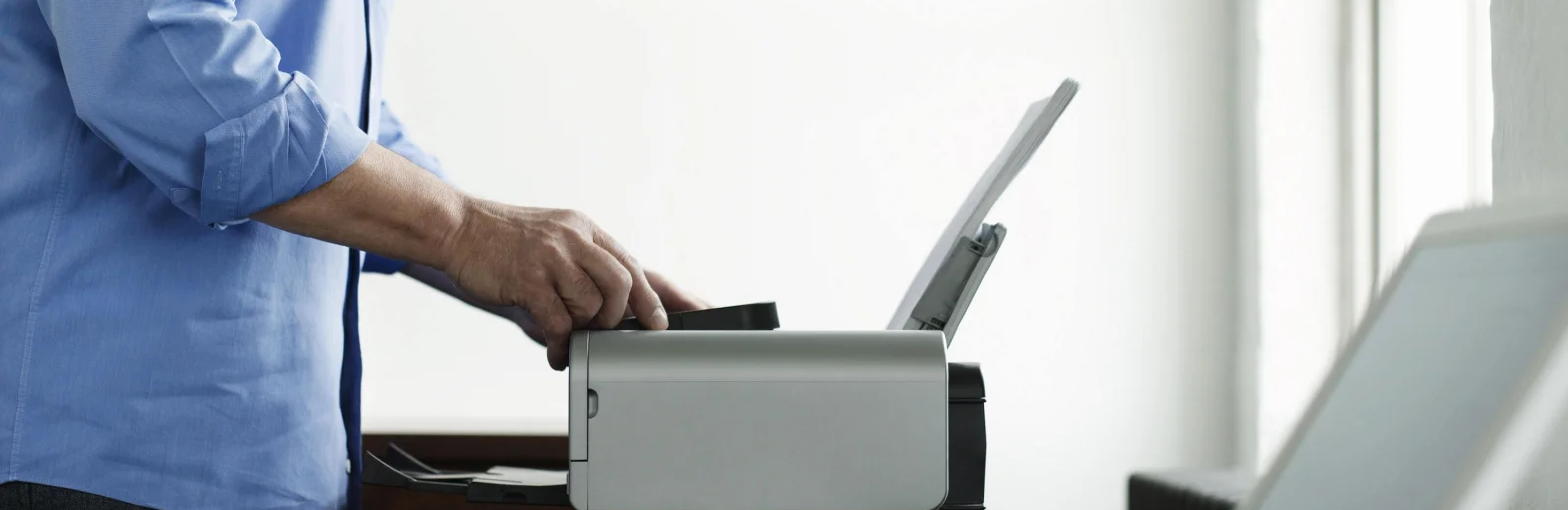 A man operates a printer on a desk, surrounded by office supplies and paperwork. 